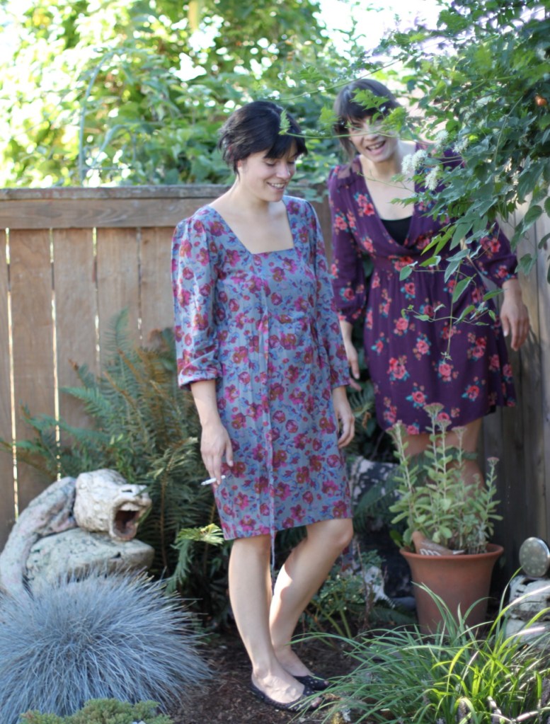 A full shot photograph of two dark haired women, both in floral patterned dresses of purple hues and shades. They stand in a garden and one woman's face is obscured by vines. One woman is holding a cigarette. They are both laughing. In the bottom left corner of the picture is a sculpture of a horse head that looks to be screaming.