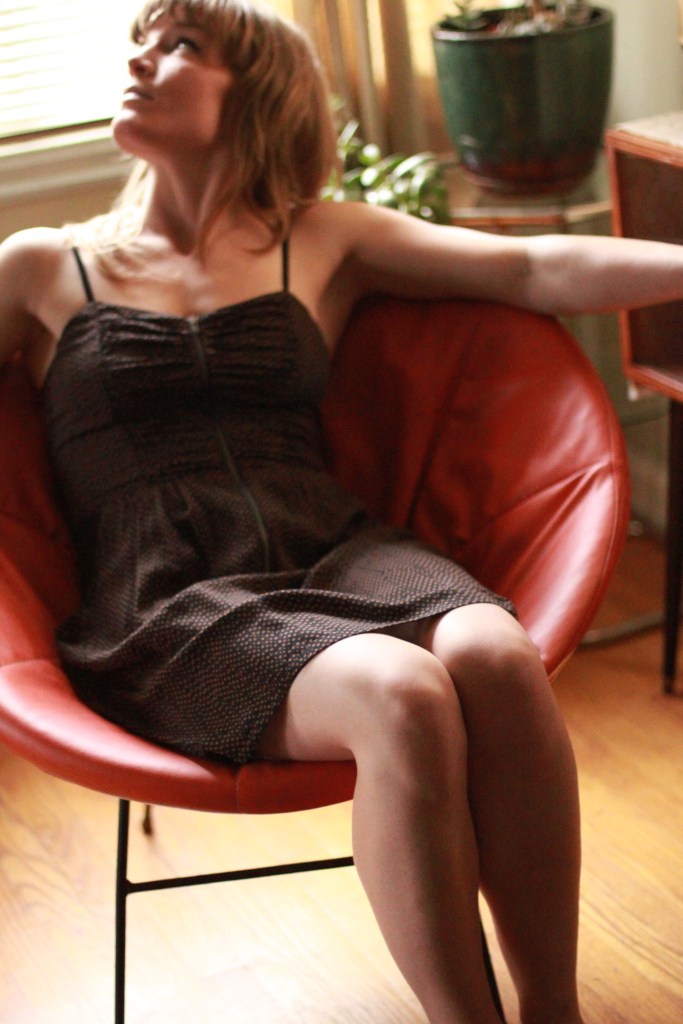 A photograph of a woman with golden blonde hair looking over her shoulder. She sits on an orange midcentury modern papasan chair with metal legs.