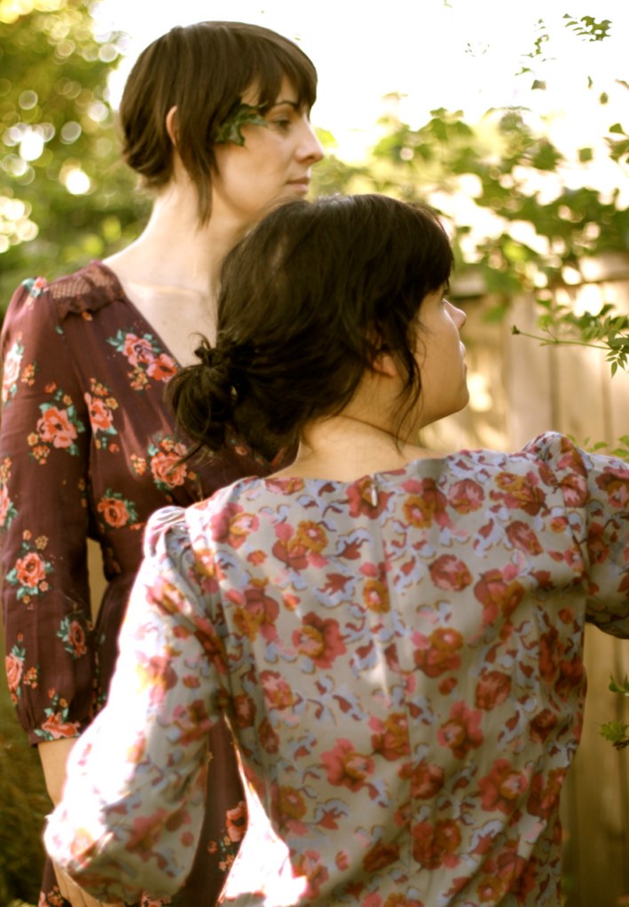 A medium close-up of two women in profile dressed in floral patterns. The women are looking at the leaves of plants.