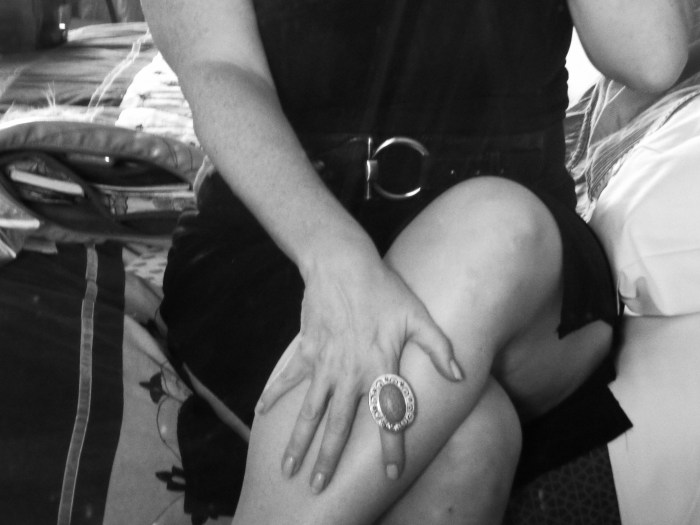 A black and white medium shot of a woman's knees and torso. Her hand is on her leg, and on her pointer finger is a large round ring. 