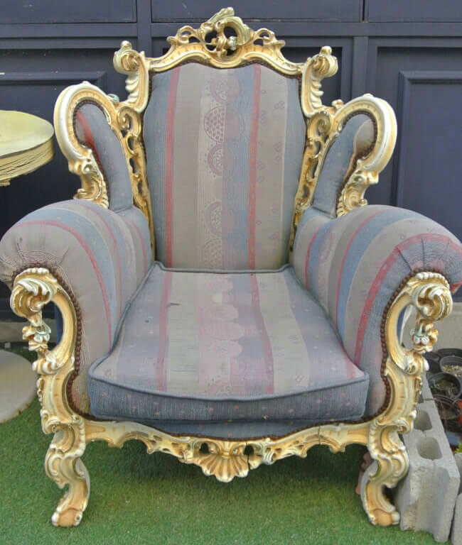 A slightly dirty old chair out side a navy blue building. The vivid stripes and geometric patterns on the Queen Anne chair in periwinkle, cobalt blue, and burgundy showcases the Queen Anne aesthetic through a delightful combination of colors and patterns. 