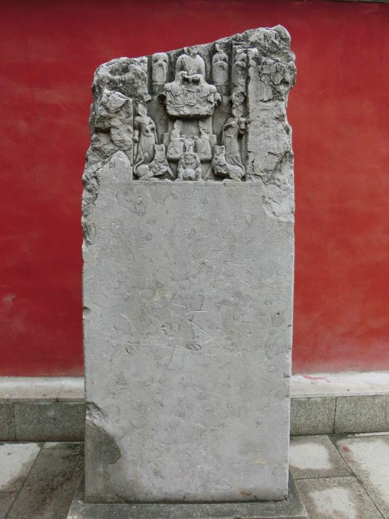 A stone tablet with mid-relief sculptures of taoist images. Set against a vibrant red wall.