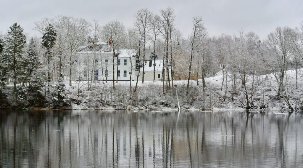 A white New England colonial style home on a snow covered bank in East Machias, Maine. The smooth Machias river reflects trees on the edge of the bank.
