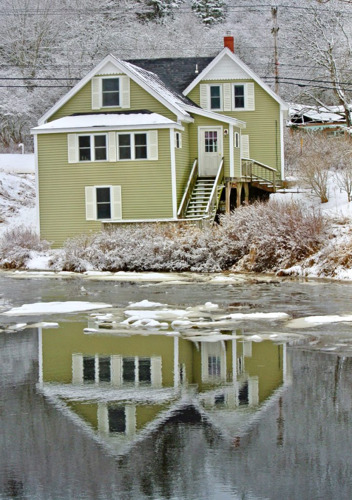  A photograph of an olive green New England house in Maine reflecting in a river with snow all around. 